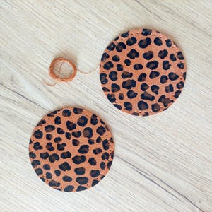 Puede incluir: Dos posavasos de cuero marrón con diseño de estampado de leopardo. Los posavasos son redondos y tienen un borde cosido.