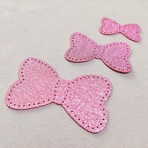 May include: Three pink glittery bow-shaped appliques. The appliques are in varying sizes, with a textured surface and stitched edges. The bows are arranged on a light-colored surface, showcasing their details.