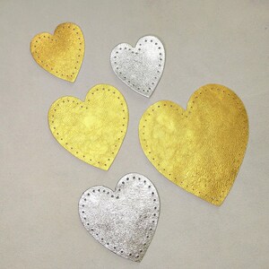 May include: Five heart-shaped leather patches in gold and silver. Each heart has a stitched border. The gold hearts have a metallic sheen, while the silver hearts have a textured appearance. The patches are scattered on a light gray surface.