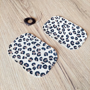 Puede incluir: Dos parches de tela beige con estampado de leopardo marrón y negro. Los parches son rectangulares y tienen un borde cosido.