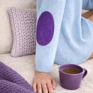 May include: A light blue long-sleeved knit jumper featuring a solid purple oval-shaped elbow patch. The jumper is paired with a purple mug and a purple knit blanket, showcasing a consistent colour palette of soft blue and deep purple tones.