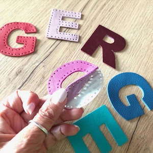 May include: A selection of leather letters in red, pink, burgundy, teal, and blue. The letter "O" is being peeled to reveal a sticky backing. The letters are stitched around the edges and are on a wooden surface.