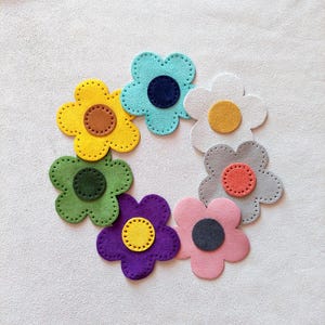 Pode incluir: Sete formas de flores de feltro dispostas em círculo. As flores são nas cores amarelo, verde, azul, branco, cinza, roxo e rosa. Cada flor tem um círculo menor no centro em uma cor contrastante.
