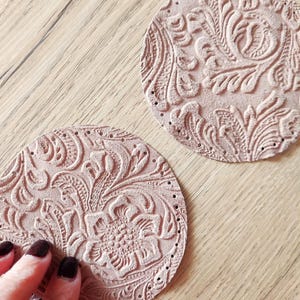 Può includere: Due sottobicchieri rotondi in pelle rosa con un design floreale in rilievo. I sottobicchieri hanno piccoli fori attorno ai bordi.