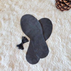 May include: A pair of dark gray, flat, shoe-shaped insoles made of a suede-like material. Each insole features a series of small, evenly spaced pre-punched holes along the entire perimeter. A small bundle of thin black cord is included nearby.