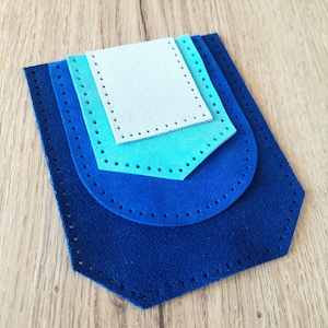 Puede incluir: Una pila de cuatro piezas de fieltro en tonos de azul y azul claro, con agujeros preperforados para coser. Las piezas están superpuestas, con una pieza superior rectangular y piezas inferiores redondeadas.