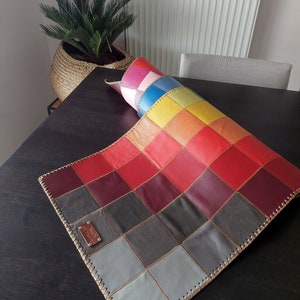 May include: A colorful leather patchwork rug with a stitched border. The rug is made up of squares of different colors, including red, orange, yellow, green, blue, and purple. The rug is rolled up and has a label that says "Genuine Leather".