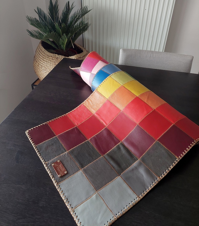 May include: A colorful leather patchwork rug with a stitched border. The rug is made up of squares of different colors, including red, orange, yellow, green, blue, and purple. The rug is rolled up and has a label that says "Genuine Leather".