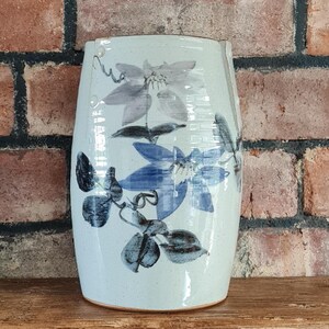 May include: A ceramic vase with a light blue background and a floral design in shades of blue and gray. The vase has a cylindrical shape with a slightly flared top and is displayed on a wooden surface.