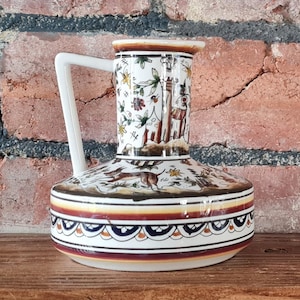 May include: A white ceramic pitcher with a brown, white, and blue floral design. The pitcher has a handle and a spout. It is sitting on a wooden surface.