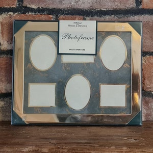 May include: A multi-aperture photo frame with a gold-colored border and a dark teal background. The frame has five openings, two rectangular and three oval. The label reads "Photofame" from Marks & Spencer.