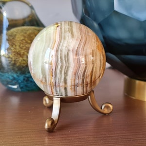 Puede incluir: Una esfera decorativa de ónix con rayas marrones, blancas y verdes. La esfera está sobre un soporte de metal dorado con tres patas.