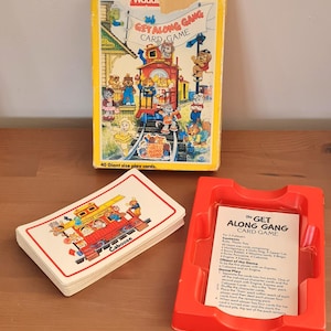 Könnte beinhalten: Das Get Along Gang Kartenspiel-Set besteht aus einer gelben Pappschachtel mit einer bunten Zugillustration, einem Kartendeck mit einem roten Caboose-Waggon und einer roten Kunststoffschale, die das gedruckte Spielanleitungsblatt enthält.