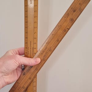 Könnte beinhalten: Zwei alte Holzlineale, eines vertikal und eines abgewinkelt, mit geätzten Markierungen und dem Text "H.O.R." sichtbar. Die Lineale sind hellbraun und scheinen alt zu sein. Die Lineale werden von einer Person gehalten.