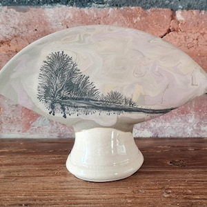 Puede incluir: Un plato de cerámica con un esmalte marmoleado blanco y rosa. El plato tiene forma de abanico y tiene una silueta en blanco y negro de árboles y un río en la parte delantera. El plato está sobre un pedestal blanco.