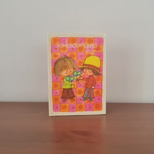 Puede incluir: Un libro vintage con una cubierta a cuadros rosa y naranja que presenta a dos niños, uno con un sombrero amarillo, intercambiando flores. El título del libro es "Somebody Cares".