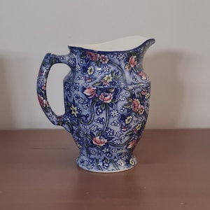May include: A blue ceramic pitcher with a floral pattern. The pitcher features a handle and a spout. The floral design includes pink and yellow flowers with green leaves. The pitcher is approximately 8 inches tall.