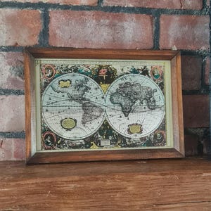 Op de afbeelding: Een antieke wereldkaart met een donkerhouten lijst. De kaart is een gedetailleerde illustratie met een vintage esthetiek, met continenten, oceanen en decoratieve elementen. De lijst staat op een houten oppervlak tegen een bakstenen muur.