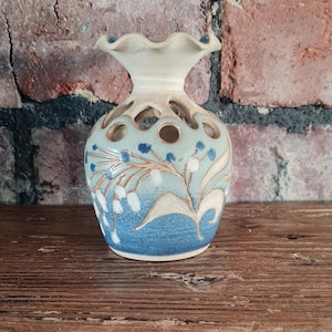 Può includere: Un vaso di ceramica con la parte superiore a balze e un corpo rotondo. Il vaso è dipinto con un motivo floreale blu e bianco. Il corpo presenta ritagli circolari. Il vaso è su una superficie di legno contro un muro di mattoni.