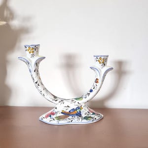 Könnte beinhalten: Weißer Kerzenständer aus Keramik mit geschwungenem Design und zwei Kerzenhaltern. Der Kerzenständer ist mit bunten Blumen- und Vogelmotiven mit blauen, gelben und grünen Akzenten verziert. Der Sockel hat eine breite, dekorative Form.