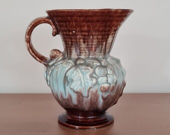 May include: A brown and blue ceramic pitcher with a handle and a decorative design of leaves and grapes.