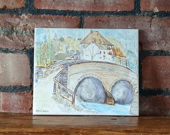 Op de afbeelding: Een aquarel schilderij van een stenen brug met gewelfde openingen over een rivier. Een gebouw met een bruin dak staat op de brug. Bomen en andere gebouwen zijn op de achtergrond. De handtekening van de kunstenaar, RECKHOLK, staat linksonder.