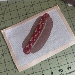 May include: A needlepoint canvas featuring a hot dog design with a brown bun, a red sausage, and yellow mustard, bordered by beige fabric tape. The canvas is positioned on a green cutting mat with a ruler showing measurements up to 11 inches.