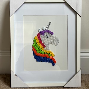 May include: A white framed picture with a unicorn made out of colorful buttons. The unicorn is outlined in white buttons and has a rainbow mane and tail.
