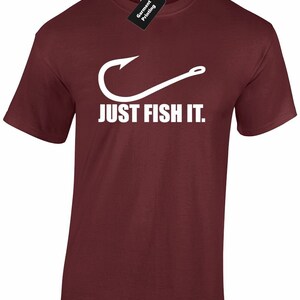 May include: Maroon t-shirt featuring a white fishing hook design and the words "JUST FISH IT." The top has a crew neck and short sleeves. A black tag with white text that says "Garment Printing" is attached to the collar.