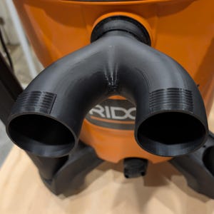 May include: Black plastic vacuum attachment with two curved nozzles. The attachment is connected to an orange RIDGID vacuum cleaner. The vacuum cleaner is on a light brown surface.