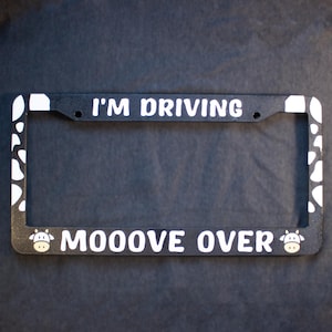 May include: Black license plate frame with white text that reads "I'M DRIVING MOOOVE OVER". The frame has a cow print design on the corners and cow graphics on each side of the text.