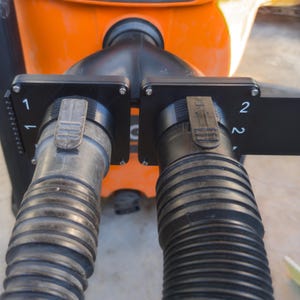 May include: Close-up of a black plastic dual hose connector with two ribbed hoses. Each hose is connected to a black rectangular plate with the numbers "1" and "2" printed on them. The background is orange.