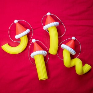 May include: Four yellow pasta-shaped Christmas ornaments, each topped with a red Santa hat with white trim and a silver string for hanging. The ornaments are arranged on a red background.