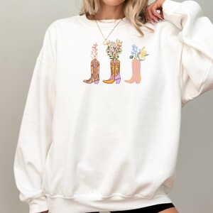 Pictured is a white Gildan crewneck sweatshirt centered on the front are three cowgirl boots in different colors of purples, pinks, and orange. Sticking out from each boot are different kinds of flowers.