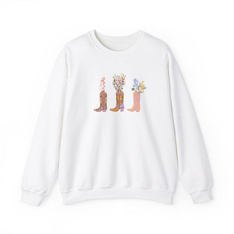 Pictured is a white Gildan crewneck sweatshirt centered on the front are three cowgirl boots in different colors of purples, pinks, and orange. Sticking out from each boot are different kinds of flowers.