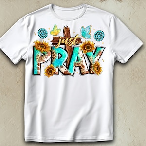 May include: White t-shirt with a turquoise and brown graphic design that says "Just Pray". The graphic includes a cross, butterflies, and sunflowers.