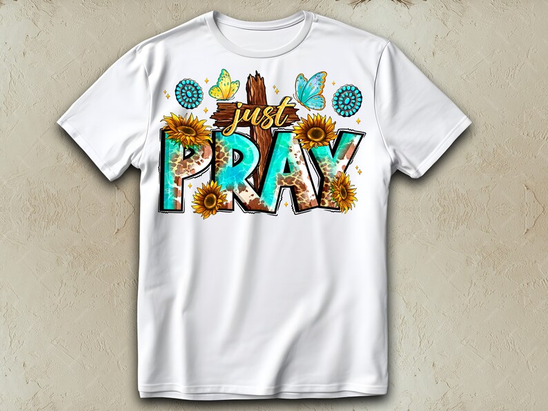 May include: White t-shirt with a turquoise and brown graphic design that says "Just Pray". The graphic includes a cross, butterflies, and sunflowers.