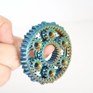 Può includere: Un ingranaggio multistrato con finitura iridescente blu e oro. L'ingranaggio ha diversi ingranaggi più piccoli all'interno del suo design. Il prodotto è tenuto su uno sfondo bianco.
