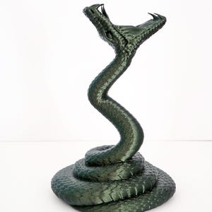 Op de afbeelding: Een donkergroene sculptuur van een slang. De kop van de slang is omhoog, de bek open, de tanden zichtbaar. Het lichaam is aan de basis opgerold en buigt omhoog. De sculptuur heeft een getextureerd oppervlak.
