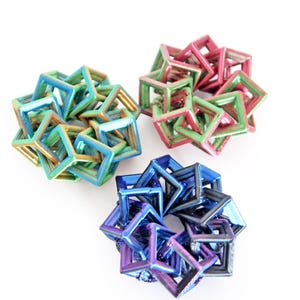 Peut inclure: Trois sculptures géométriques en forme de cubes, aux couleurs irisées. L'une est bleue et violette, une autre verte et dorée, et la troisième rose et verte. Elles sont disposées sur un fond blanc.