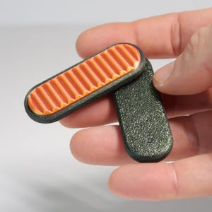 Puede incluir: Un pequeño objeto rectangular con bisagras, con un interior naranja acanalado y un exterior oscuro y moteado. El objeto se sostiene en la mano de una persona sobre un fondo blanco. El objeto parece estar hecho de plástico.