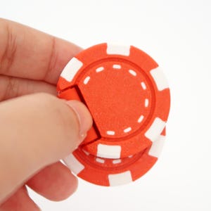 Puede incluir: Primer plano de dos fichas de póquer rojas y blancas. Las fichas son circulares, con una base roja y detalles blancos rectangulares en el borde. Una mano sostiene las fichas. Las fichas se utilizan para juegos.