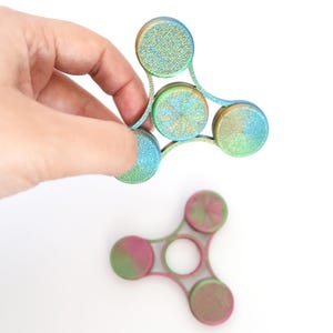 Peut inclure: Deux hand spinners colorés sur fond blanc. L'un est tenu par une main, avec un dégradé de bleu, vert et or. L'autre est rose et vert. Les spinners ont un design à trois lobes avec des bords arrondis.