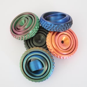 Op de afbeelding: Een verzameling kleurrijke, metalen fidget spinners in groen, blauw en oranje tinten. Elke spinner heeft een uniek ontwerp, met getextureerde randen en concentrische cirkels. De spinners zijn gerangschikt op een witte achtergrond. Afmetingen in cm.