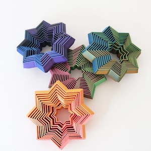 Peut inclure: Collection de six objets décoratifs en forme d'étoile multicolores. Chaque étoile est composée de formes géométriques superposées dans des tons de violet, bleu, vert, orange et rose. Les objets sont disposés sur une surface blanche.