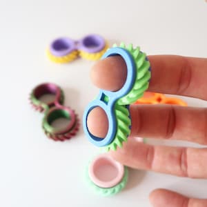 Pode incluir: Uma coleção de anéis coloridos impressos em 3D em vários designs. Um anel é segurado, mostrando uma moldura azul com bordas texturizadas verdes e azuis. Outros anéis no fundo apresentam tons roxos, rosa e amarelo. Os anéis estão em um fundo branco.