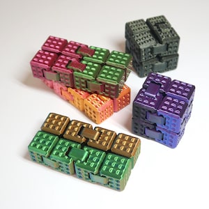 Può includere: Una collezione di colorati giocattoli a cubo ad incastro nei toni del rosa, verde, arancione e viola. I giocattoli sono disposti in varie configurazioni, alcuni come lunghe catene e altri come cubi. Lo sfondo è bianco.