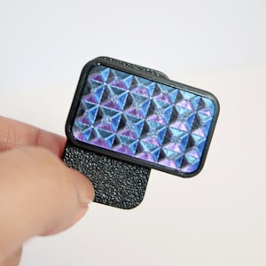Può includere: Un supporto nero per telefono con base strutturata e un motivo geometrico iridescente nei toni del blu, viola e argento. Il supporto è tenuto su uno sfondo bianco.