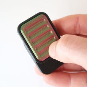 Op de afbeelding: Een klein, rechthoekig zwart object met een geribbelde, veelkleurige bovenkant, vastgehouden in een hand. De bovenkant heeft afwisselend groene en roze strepen. Het object heeft afgeronde hoeken en een gladde, matte afwerking.