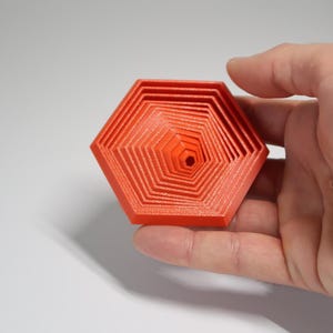 Può includere: Un esagono nidificato arancione stampato in 3D. L'oggetto, tenuto in mano, presenta una serie di forme esagonali decrescenti l'una dentro l'altra, creando un effetto a strati. Il design è geometrico e moderno.